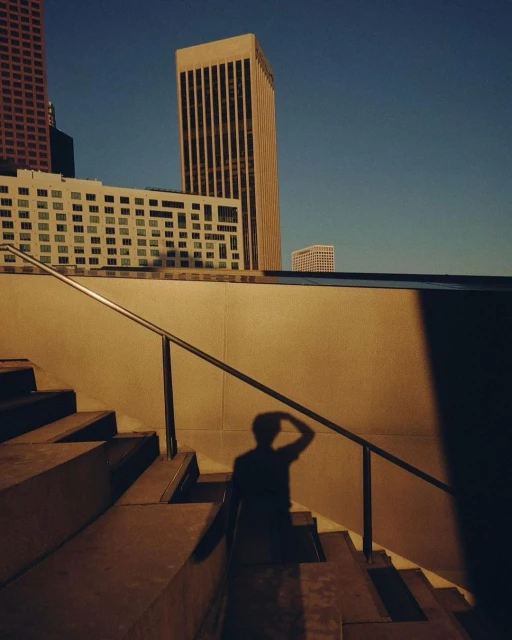 Low-angle view of a person's shadow on outdoor steps, with modern buildings in the background and a clear blue sky above.