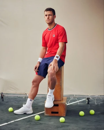 Man in red FILA shirt and blue shorts sits on wooden boxes near tennis balls in studio setting for fitness or sport product shoot.