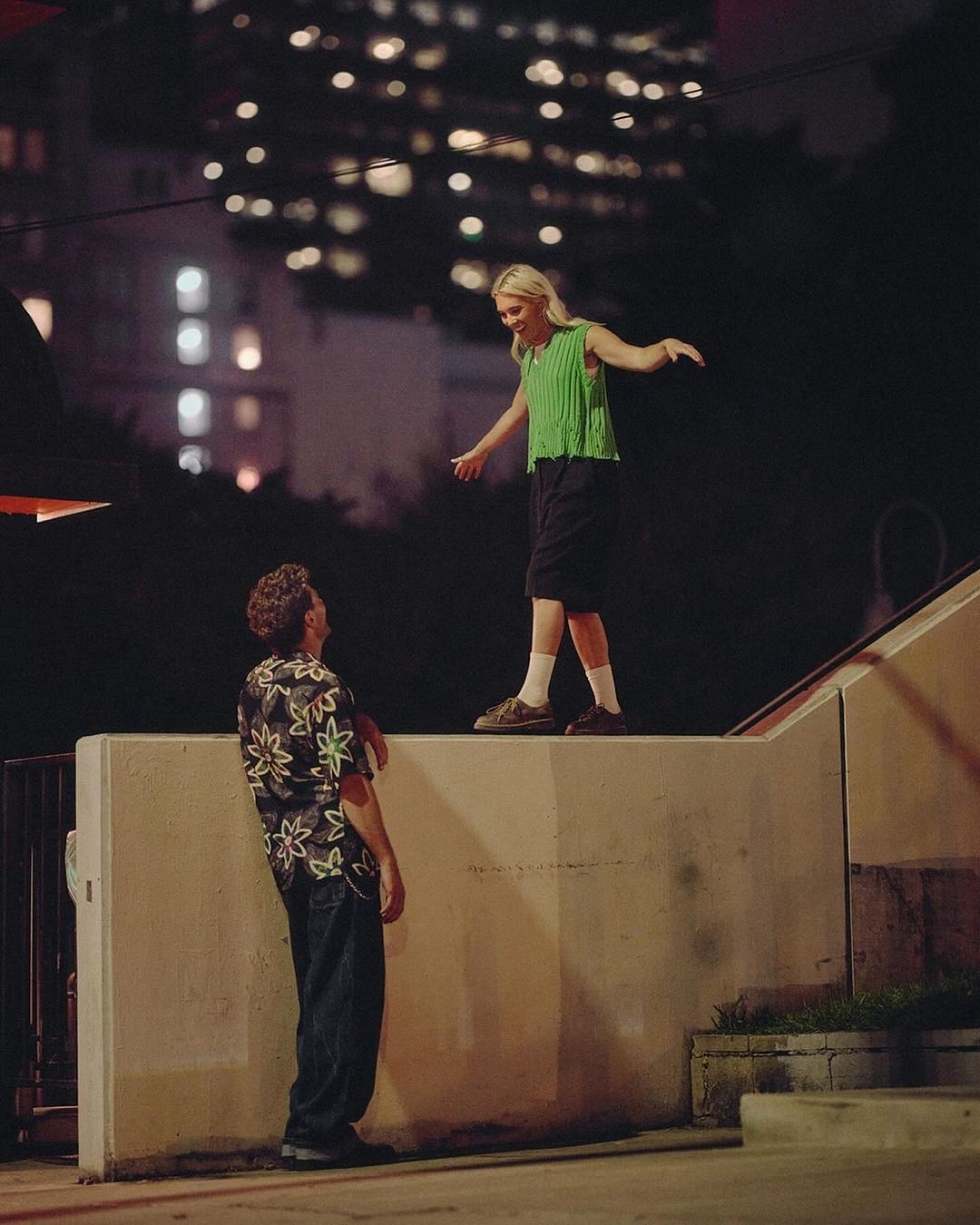 A woman balances on a wall as a man looks up at her in an urban setting at night with buildings and lights in the background.