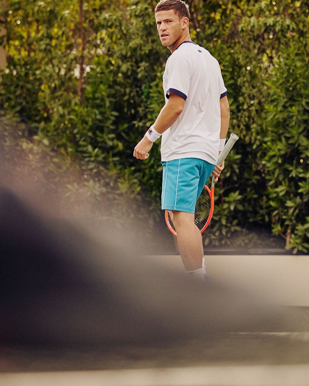 Athletic man in tennis attire with racket on outdoor court, looking over shoulder, captured in medium shot with natural light.