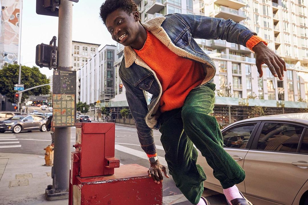 Young man in motion on city street, wearing orange sweater, denim jacket and green corduroy pants, with buildings in background.