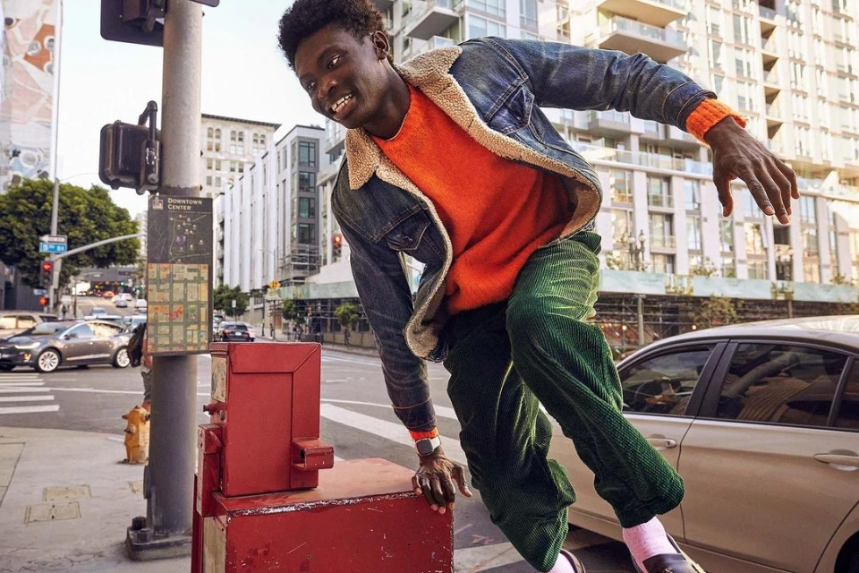 Young man in motion on city street, wearing orange sweater, denim jacket and green corduroy pants, with buildings in background.