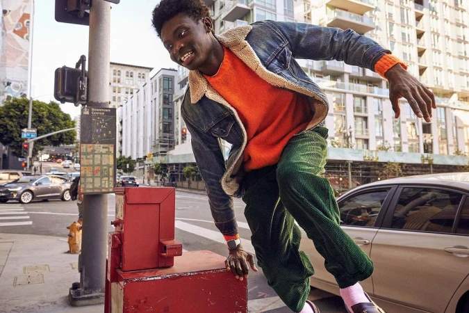 Young man in motion on city street, wearing orange sweater, denim jacket and green corduroy pants, with buildings in background.