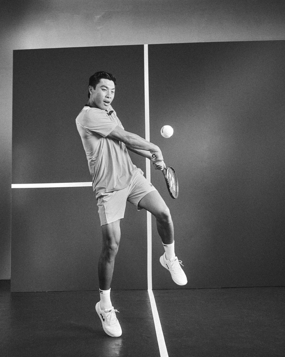 Monochrome shot of a tennis player hitting a ball with a racket indoors, wearing athletic gear, against a dark background with lines.