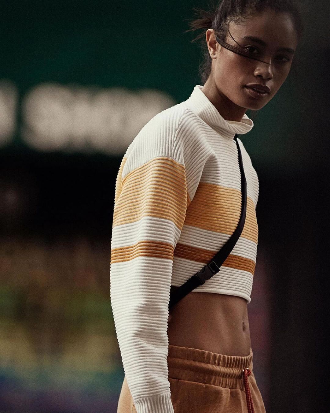 An adult woman in a crop top and sweatpants poses indoors against a dark backdrop, showcasing fashion and athletic wear.