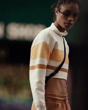 An adult woman in a crop top and sweatpants poses indoors against a dark backdrop, showcasing fashion and athletic wear.