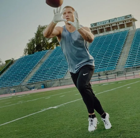 An athlete in athletic wear catches a football on the green field in front of a stadium on a sunny day, medium shot, landscape orientation.