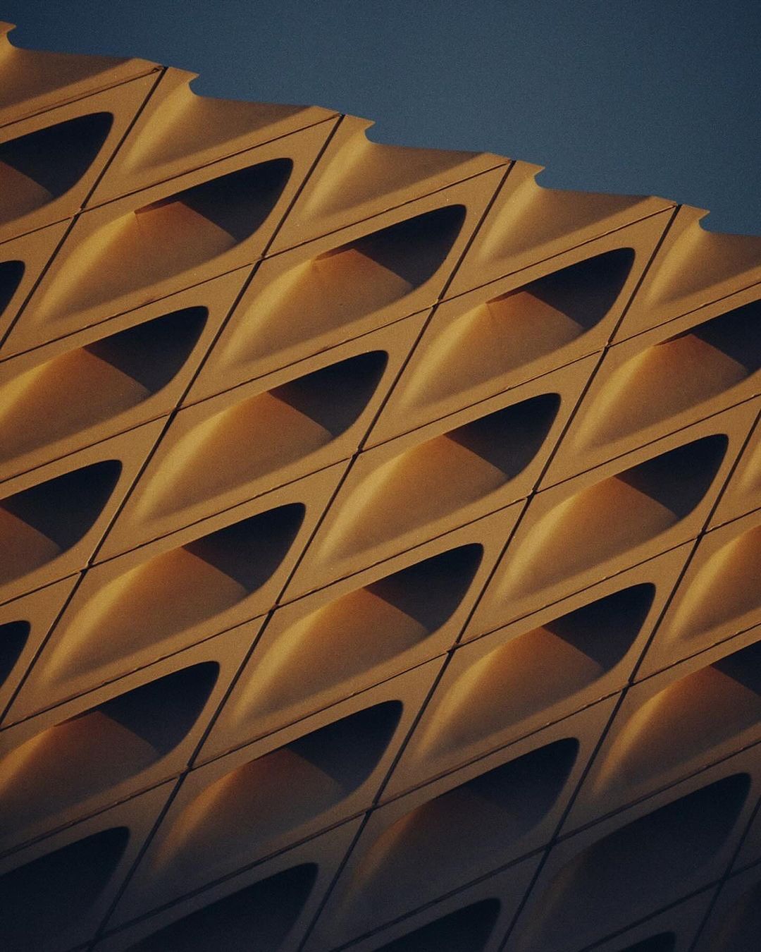 Architectural shot showcasing a building facade with a geometric pattern of repeating, concave shapes casting shadows, against a blue sky.