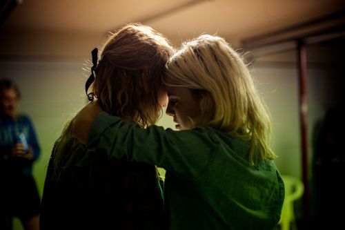 Two women with blonde hair are shown indoors in a close embrace. They are wearing dark green tops, and the background is slightly blurred.