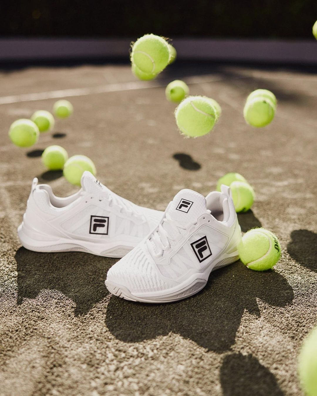 White Fila tennis shoes with scattered tennis balls on an outdoor court surface in soft natural light, product detail, sport