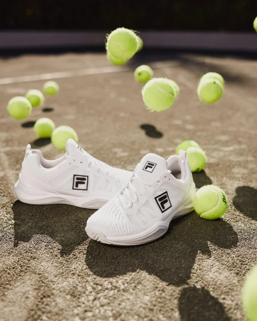 White Fila tennis shoes with scattered tennis balls on an outdoor court surface in soft natural light, product detail, sport