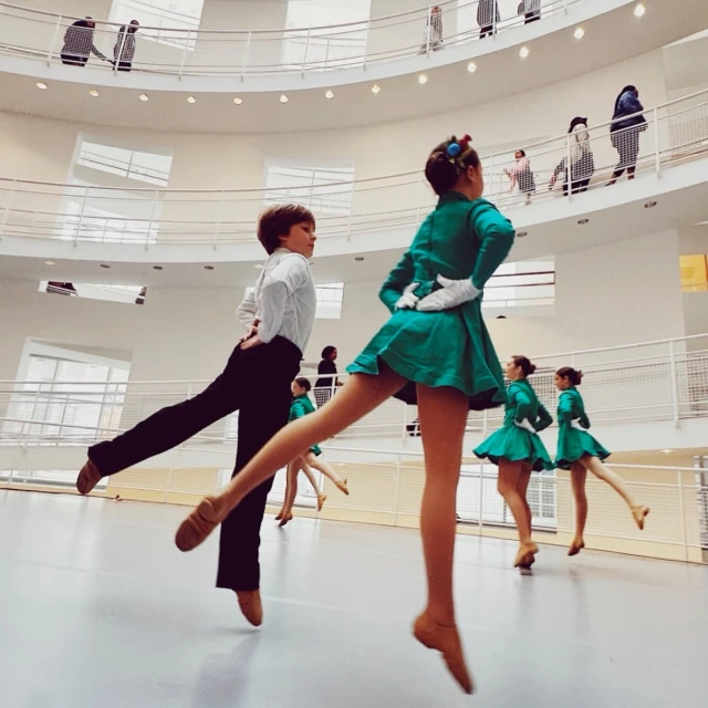 Group of young ballet dancers, including a boy, performing in a modern, light-filled architectural space with onlookers on upper levels.