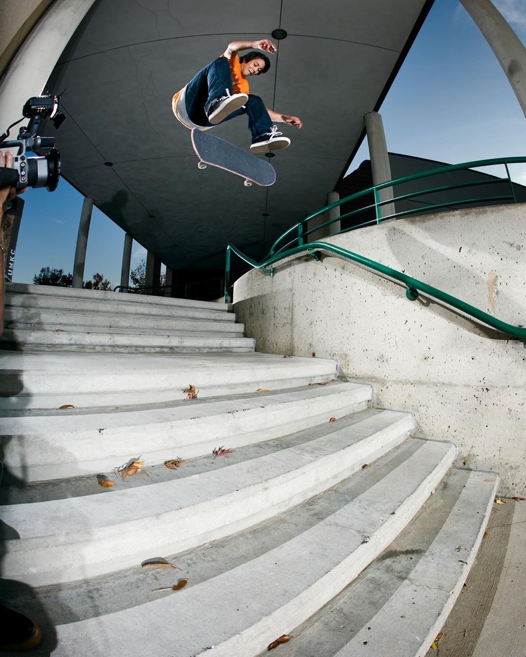 An adult male skateboarder performs a trick over concrete stairs in an outdoor setting, captured at eye-level with a blue sky backdrop.