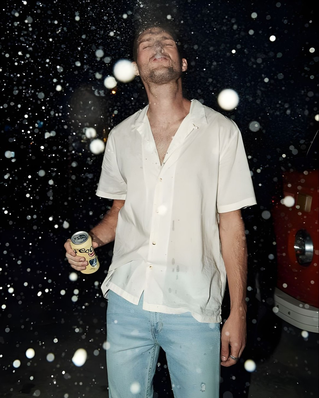 Adult male stands outside in the dark, illuminated by lights, holding a canned beverage as liquid sprays over him and nearby.