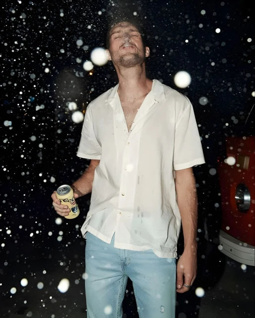 Adult male stands outside in the dark, illuminated by lights, holding a canned beverage as liquid sprays over him and nearby.