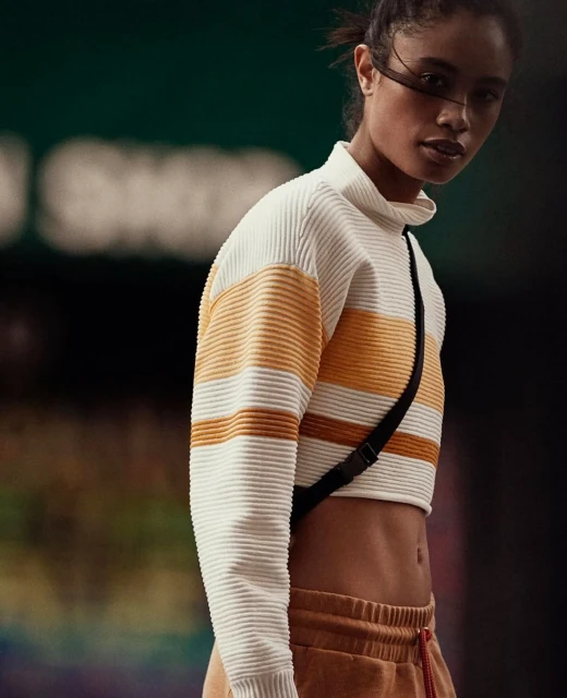 A woman poses in a cream-and-orange striped, short-sleeved sweater and sweatpants against a blurred, dark green and black backdrop.