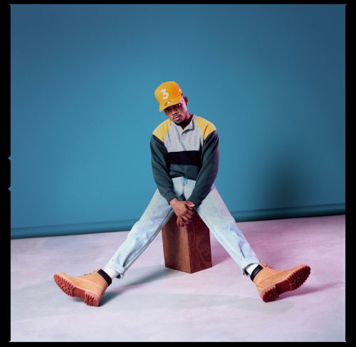 Portrait of a young adult male with light-wash jeans, a polo shirt, work boots, and a baseball cap, sitting on a wooden cube in a studio.