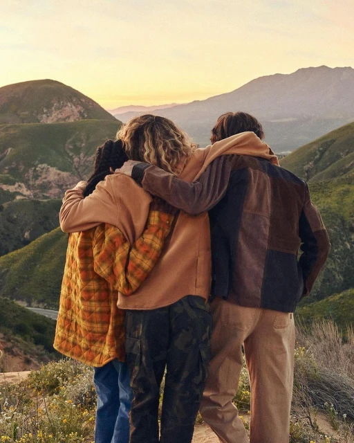 Three people embrace, standing shoulder-to-shoulder overlooking scenic mountain landscape at dusk. Friends bonding in nature.