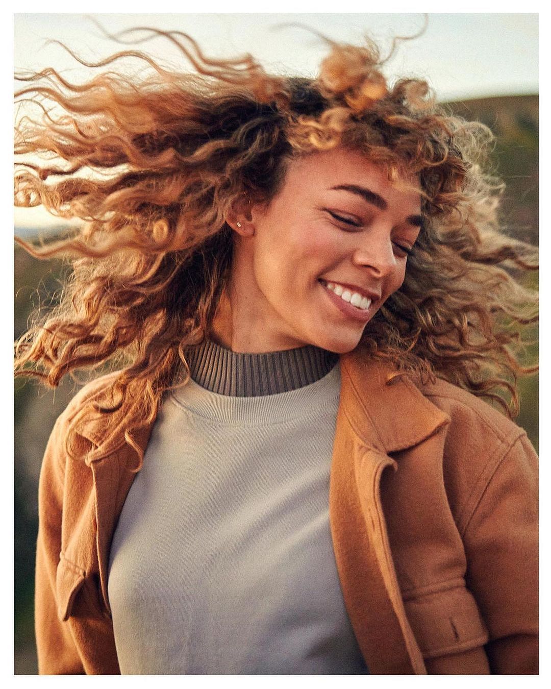 Smiling woman with curly blonde hair blowing in the wind, wearing a grey mock neck top and camel jacket outdoors.