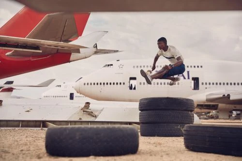 Athletic Black man jumps over tires in an outdoor plane graveyard; action shot with airplane tails in the background on a cloudy day.