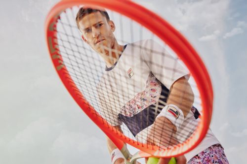 Male tennis player holds a red tennis racket up to the camera in an outdoor shot with a light sky, wearing floral athletic apparel.