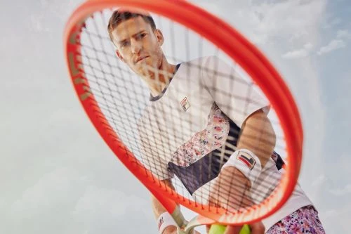 Male tennis player holds a red tennis racket up to the camera in an outdoor shot with a light sky, wearing floral athletic apparel.