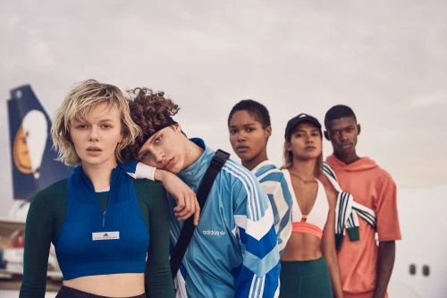Five people in athletic wear stand in front of an airplane. Two women and three men in sport jackets, tank tops, and matching pants.