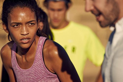 Close-up of a sweaty woman in athletic wear, with two men in the background wearing casual t-shirts in an outdoor setting.