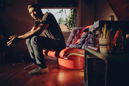 Indoor portrait of a man sitting on an orange vinyl bench seat near a window, with indirect light and a cluttered shelving unit.
