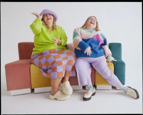 Two smiling plus-size women sit on a colorful couch in a studio, wearing a green top, patchwork skirt, patterned sweater, and stylish shoes.