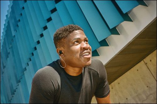 Black man wearing a dark athletic t-shirt with wired earphones looking upward against a turquoise paneled architectural wall