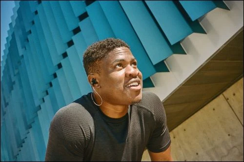 Black man wearing a dark athletic t-shirt with wired earphones looking upward against a turquoise paneled architectural wall