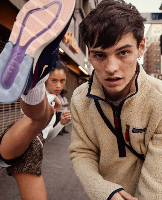 Two people pose in an urban setting. One is mid-kick, wearing patterned shorts and a sneaker; the other wears a fleece jacket.