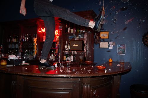 Person in black jeans and shoes kicking glass on a bar, with liquor bottles and a neon sign in the background.