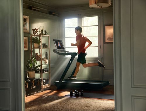 Adult male running on a treadmill inside a living room next to a window. Dumbbells on the floor near an antique trunk.