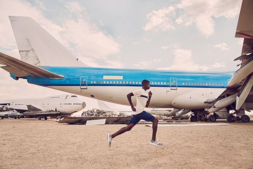 A man sprints across a sandy landscape, his form silhouetted against the backdrop of vintage airplanes under a partly cloudy sky.