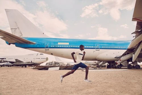 A man sprints across a sandy landscape, his form silhouetted against the backdrop of vintage airplanes under a partly cloudy sky.