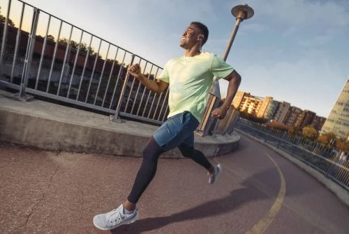 Active African American man running on curved city bridge with buildings in background on a bright, sunny day in athletic gear.