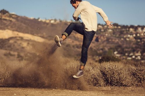 Athletic adult male leaps dynamically on a sandy path, kicking up dust and grass. The background shows rolling hills and blue sky.