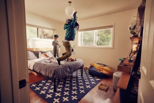 Two children are in a bright bedroom. One is jumping on a bed. The other stands and holds a toy. There is a window in the background.