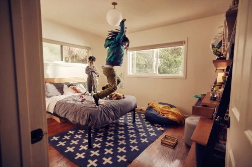 Two children are in a bright bedroom. One is jumping on a bed. The other stands and holds a toy. There is a window in the background.