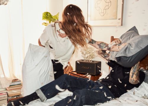 Two people having a pillow fight in a brightly lit bedroom with white walls, hardwood floors, and a bed covered in white bedding.