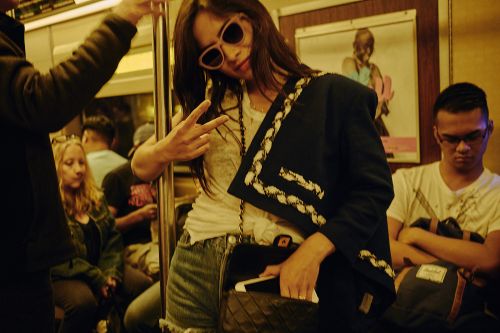 Young woman in denim shorts, white t-shirt, sunglasses, and black jacket flashing peace sign on subway with passengers in background.