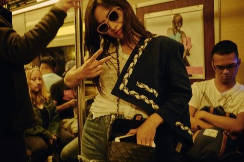 Young woman in denim shorts, white t-shirt, sunglasses, and black jacket flashing peace sign on subway with passengers in background.