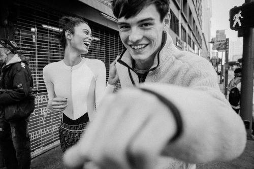 Monochrome shot of a smiling man in a fleece jacket pointing at the camera on a city street with a laughing woman and buildings in the background.