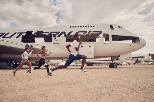 Three athletic adults running outdoors in front of a decommissioned Southern Air passenger airplane on a dirt ground under a cloudy sky.