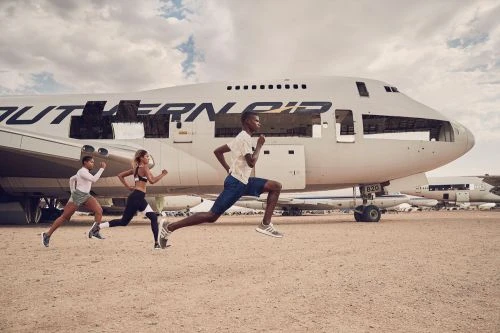 Three athletic adults running outdoors in front of a decommissioned Southern Air passenger airplane on a dirt ground under a cloudy sky.