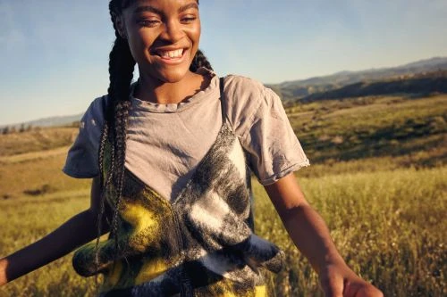 Smiling black woman in a plaid vest and grey t-shirt outdoors in a grassy meadow under a clear sky.