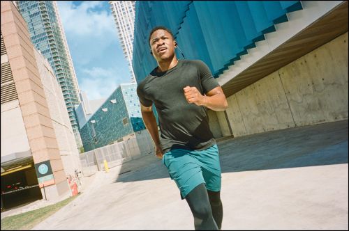 African-American male athlete running in teal shorts and a dark T-shirt between tall buildings on a sunny day in an urban setting.
