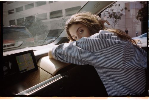 Adult female with long brown hair leans on the dashboard of a car, wearing a striped shirt, with urban buildings visible through the windshield.